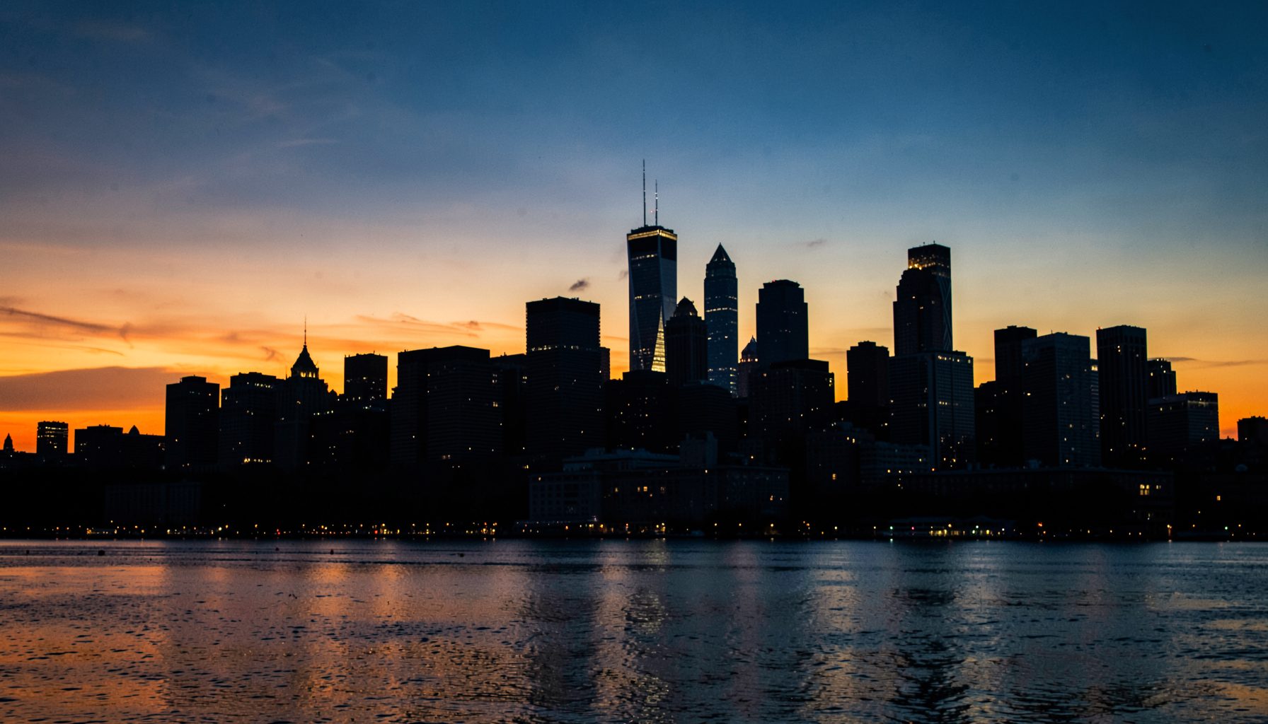 A wide cityscape of Chicago skyline at dusk, with warm amber and deep blue tones across the sky, the dark silhouettes of skyscrapers reflecting soft light off Lake Michigan in the foreground.