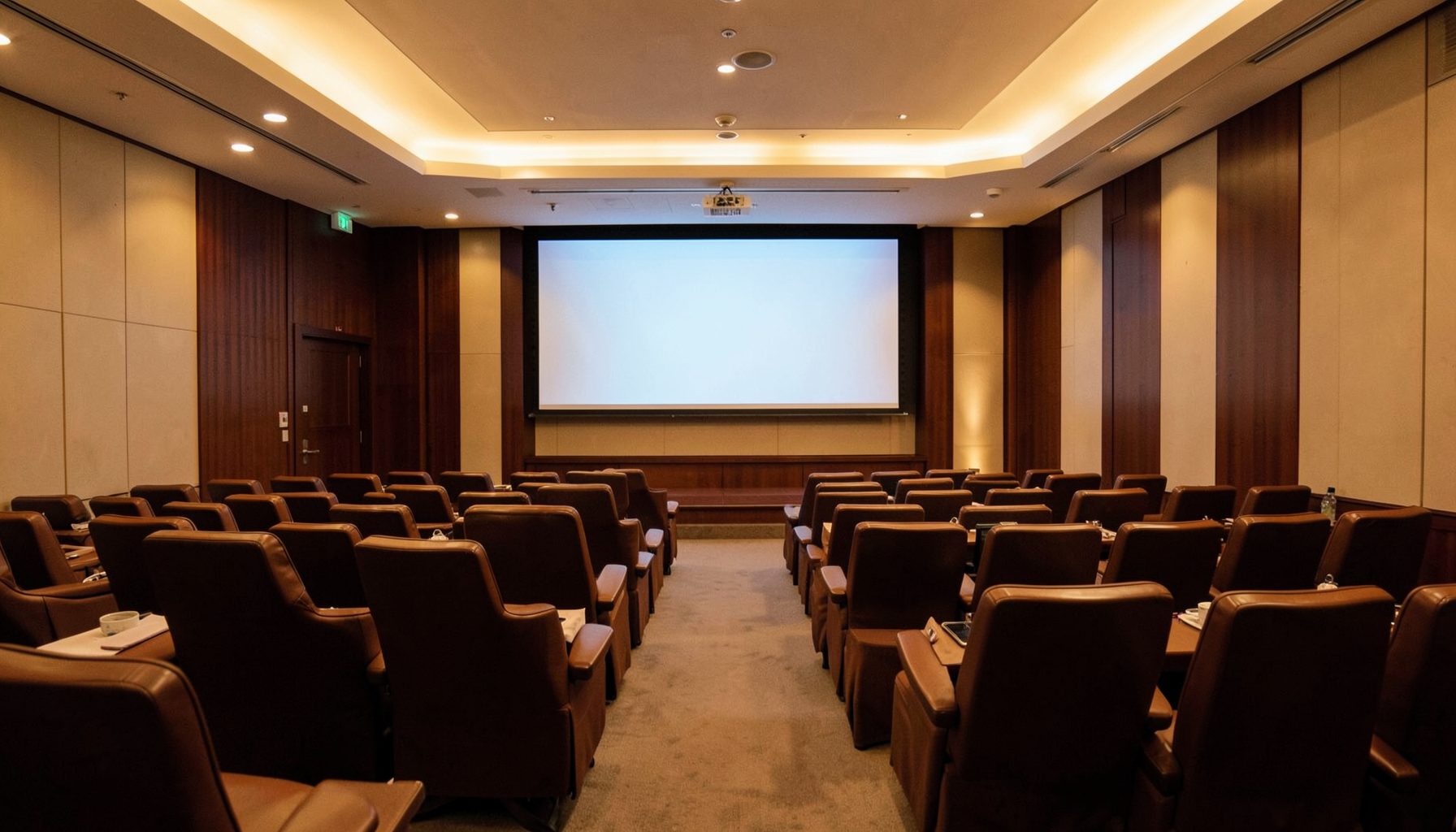 Warmly lit conference hall interior with rows of seating and a large presentation screen, deep brown and cream tones, professional atmosphere